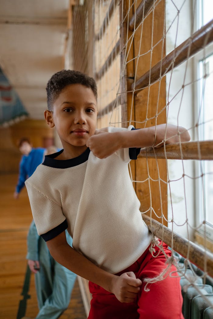 A young boy leans on a net in a gymnasium, wearing a white shirt and red pants.
